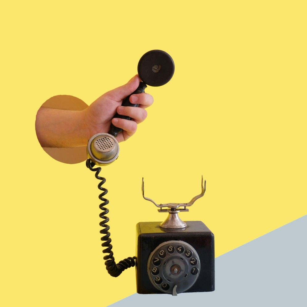 Hand holding the handset of a black rotary telephone against a yellow background